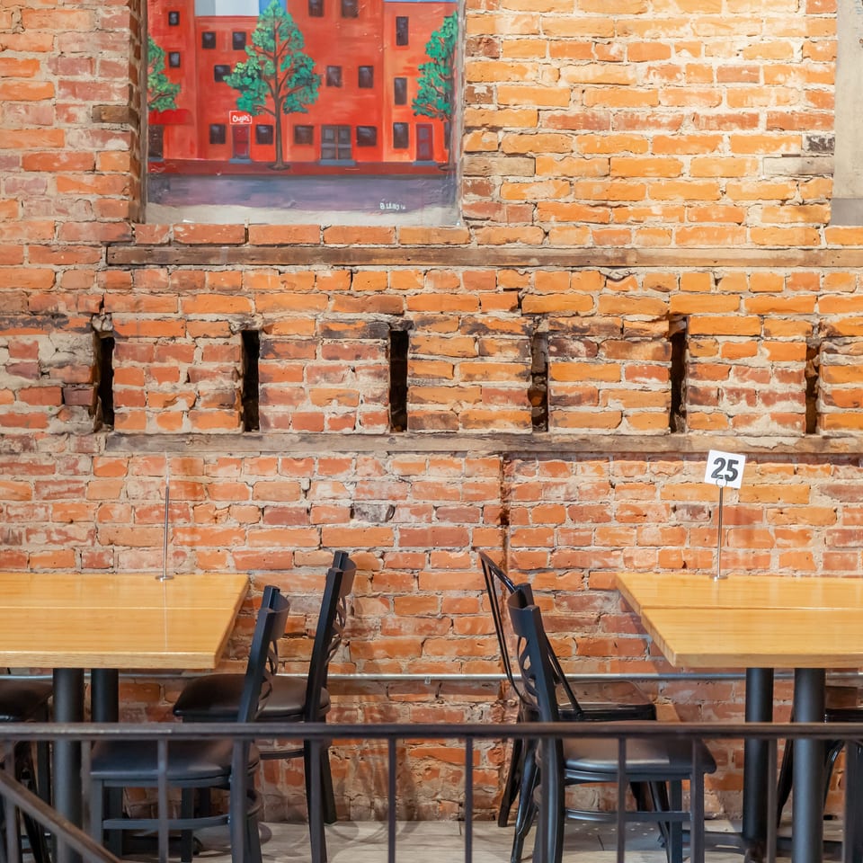 Two empty wooden tables with black chairs against a rustic brick wall with a painting.