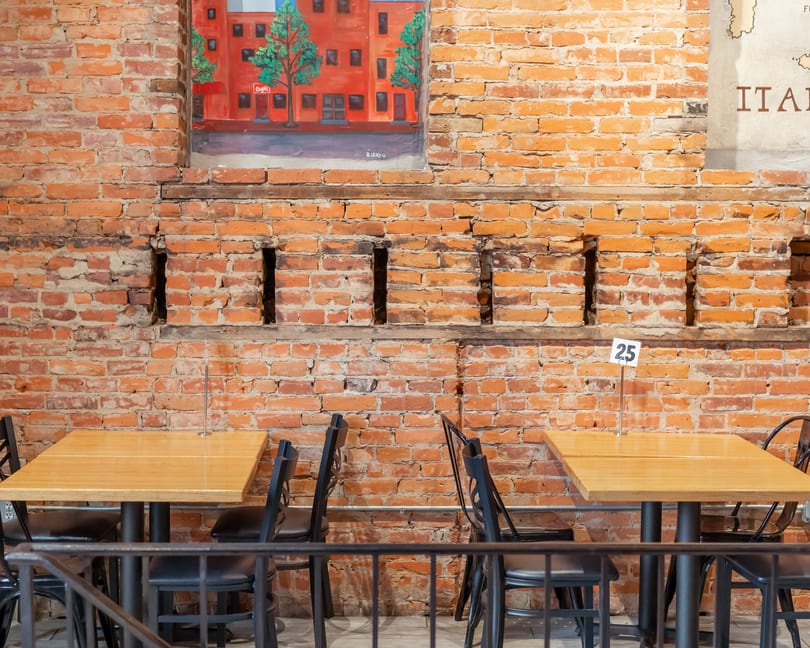 Two empty wooden tables with black chairs against a rustic brick wall with a painting.