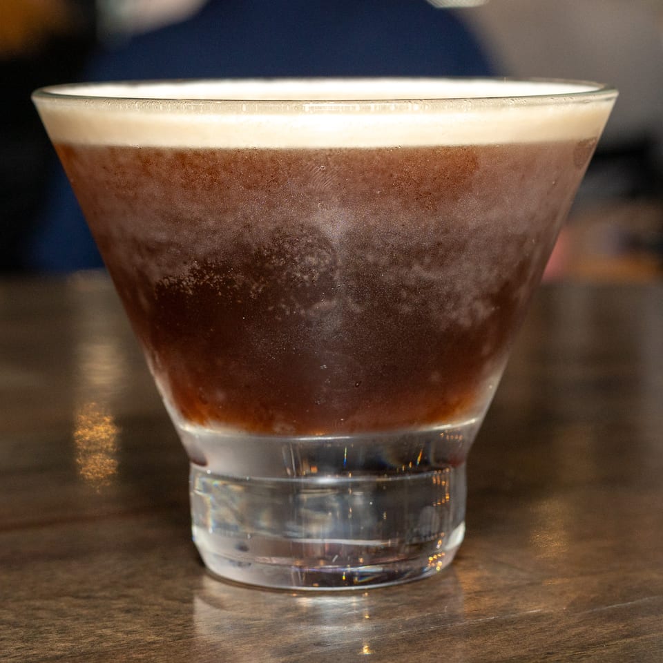 A dark, frothy espresso martini in a chilled martini glass sits on a wooden table.