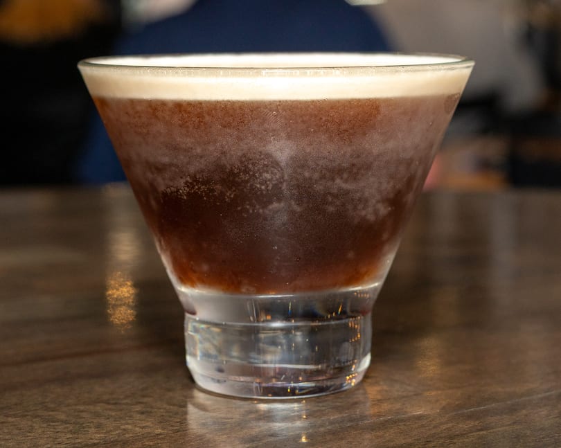 A dark, frothy espresso martini in a chilled martini glass sits on a wooden table.