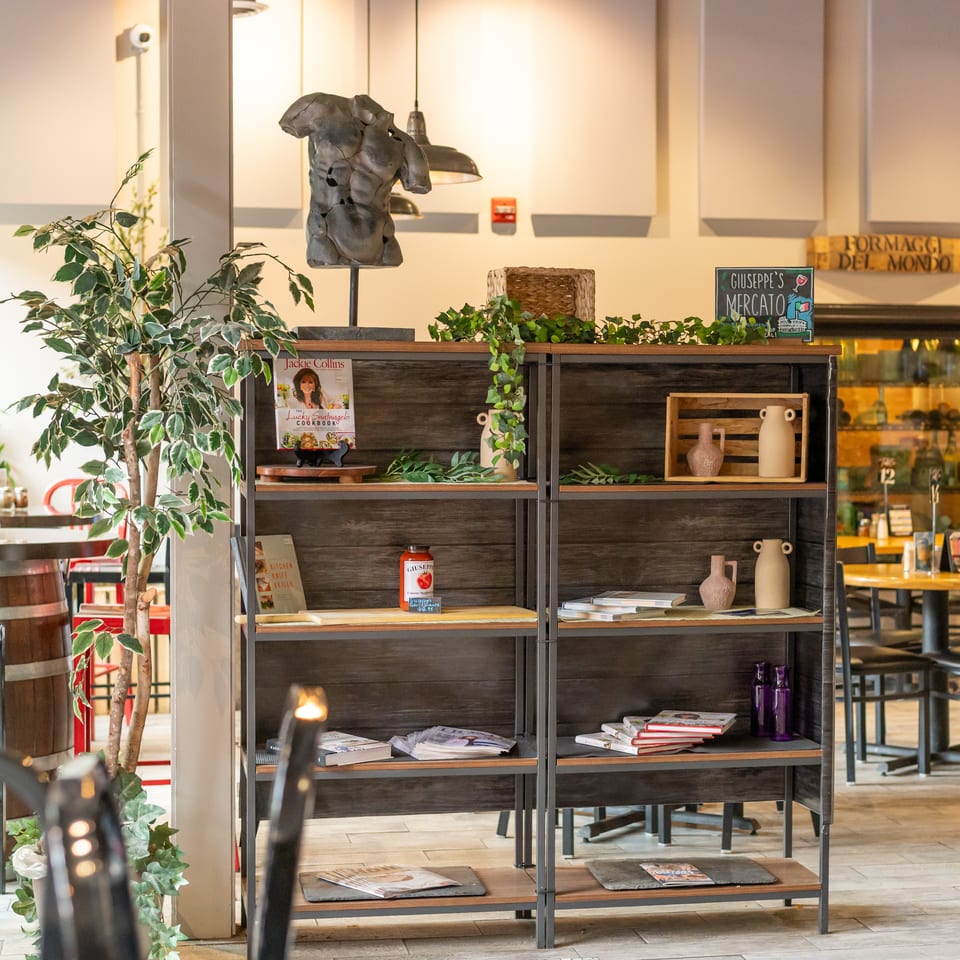 A wooden bookshelf with decor, cookbooks, and a tomato sauce jar, with restaurant seating in the background.