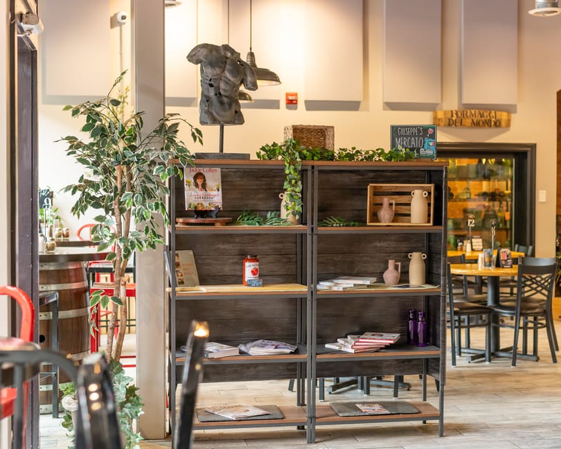 A wooden bookshelf with decor, cookbooks, and a tomato sauce jar, with restaurant seating in the background.
