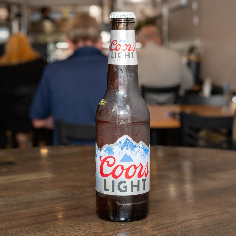 A cold bottle of Coors Light beer sits on a wooden table in a restaurant.