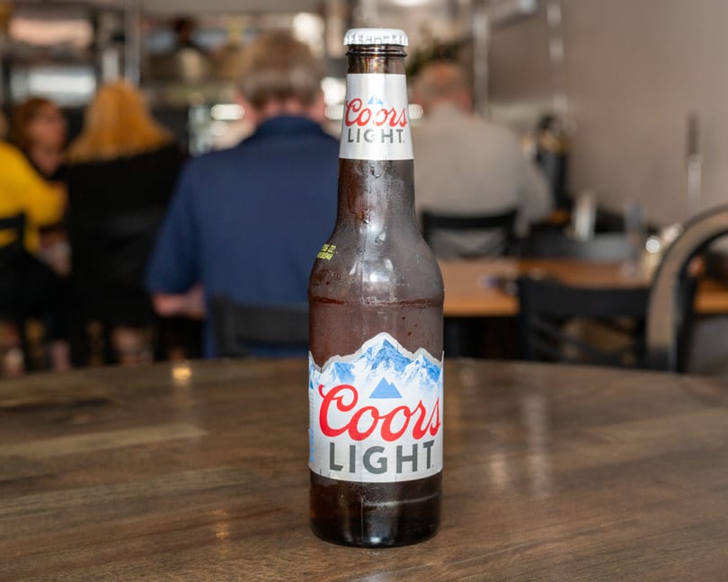 A cold bottle of Coors Light beer sits on a wooden table in a restaurant.