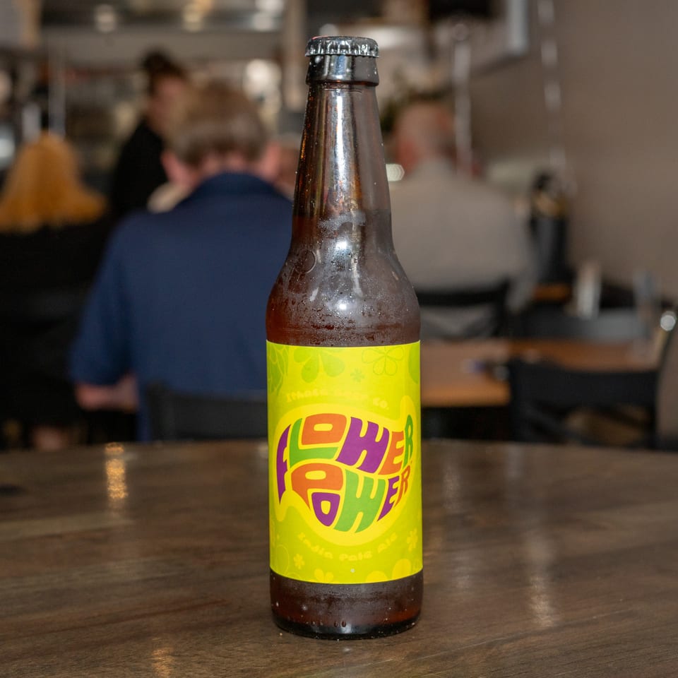 A bottle of "Flower Power" beer on a wooden table with blurred restaurant patrons in the background.