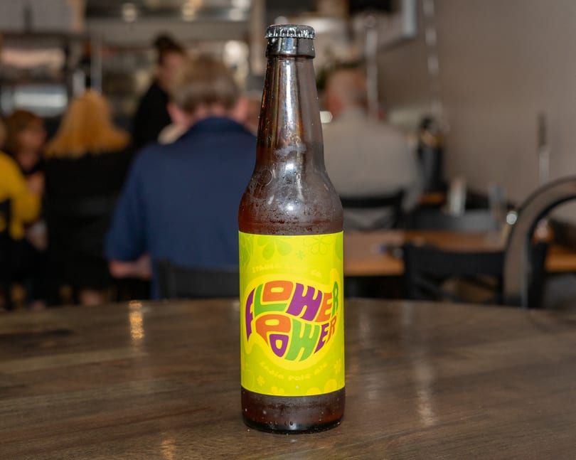 A bottle of "Flower Power" beer on a wooden table with blurred restaurant patrons in the background.