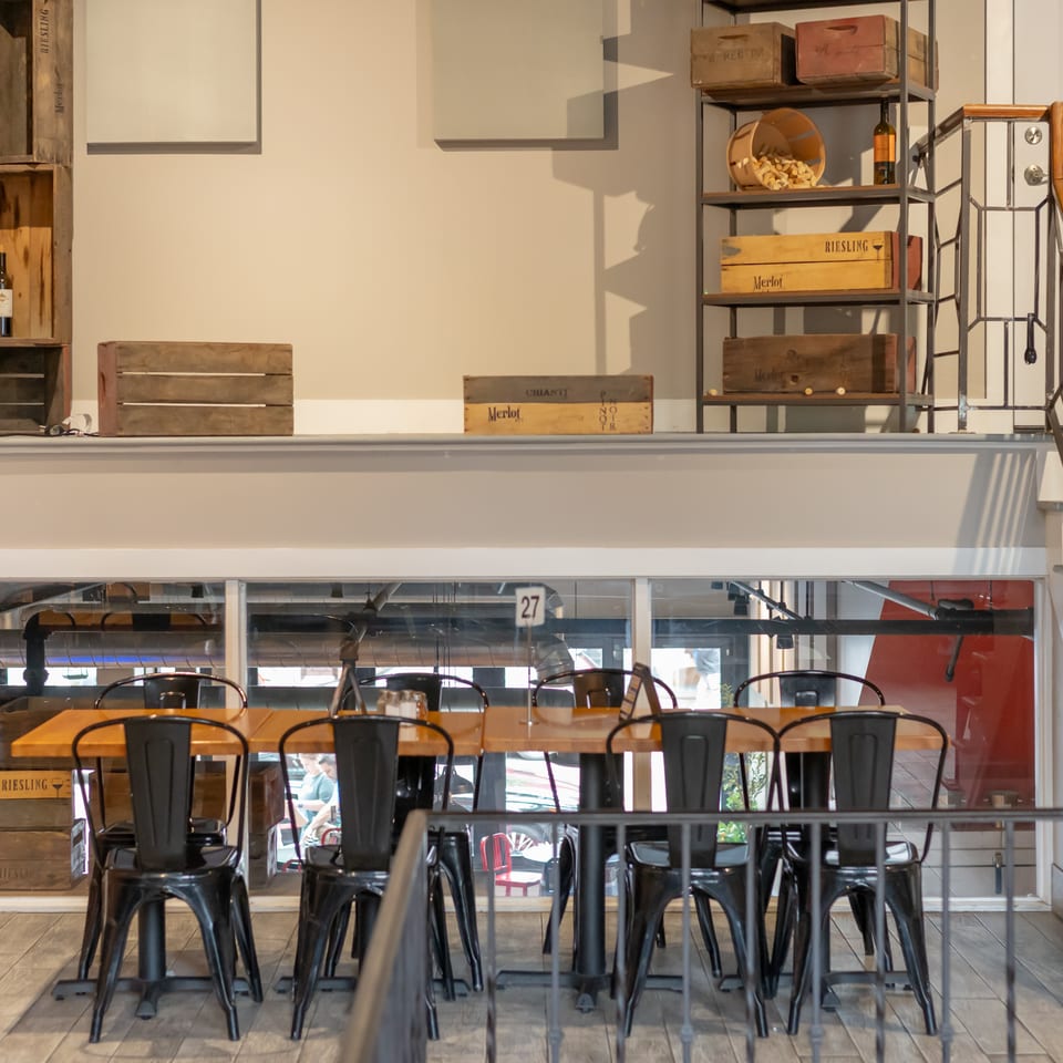 A restaurant interior with a long wooden table and black chairs, wine crates, and a staircase.