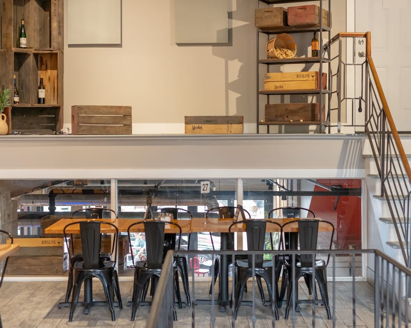 A restaurant interior with a long wooden table and black chairs, wine crates, and a staircase.