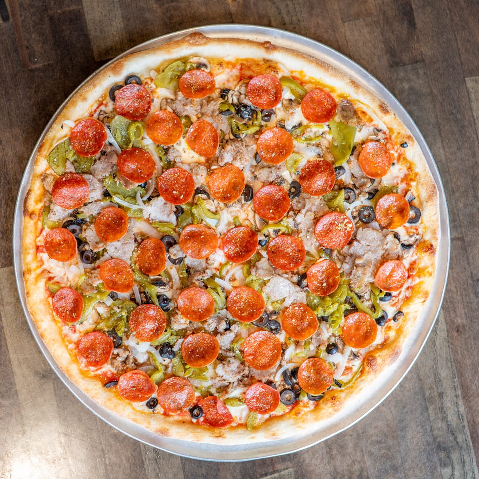 A whole pizza with pepperoni, sausage, green peppers, black olives, and onions on a wooden table.