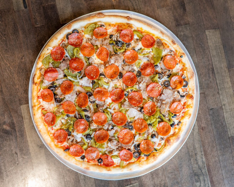 A whole pizza with pepperoni, sausage, green peppers, black olives, and onions on a wooden table.