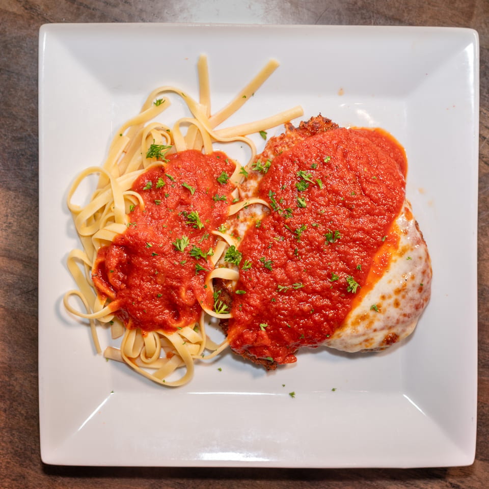 Chicken parmesan with melted cheese and red sauce, served with fettuccine pasta on a white plate.