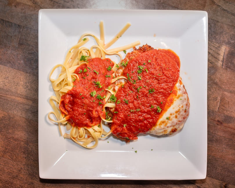 Chicken parmesan with melted cheese and red sauce, served with fettuccine pasta on a white plate.