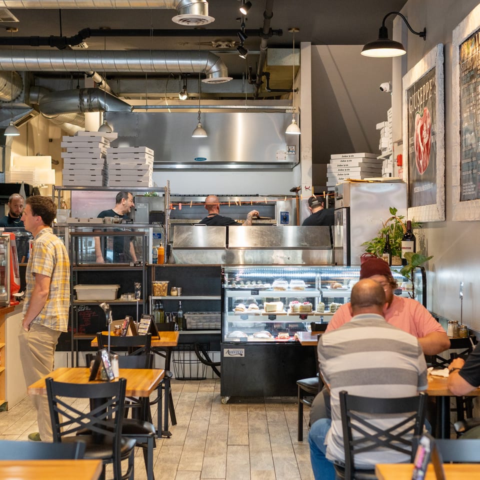 Busy pizzeria interior with customers dining, chefs working, and display cases of pastries and sauces.