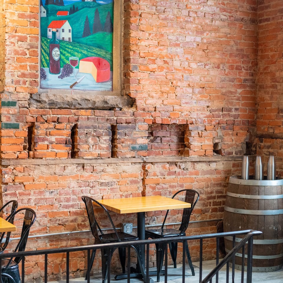 A rustic restaurant interior with exposed brick walls, a wine-themed painting, and small tables.