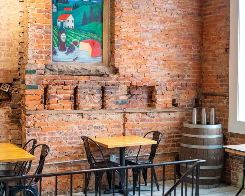 A rustic restaurant interior with exposed brick walls, a wine-themed painting, and small tables.