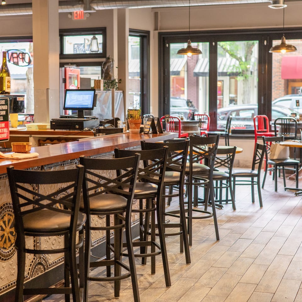 Interior of a restaurant with a long bar, bar stools, and dining tables with chairs near large windows.