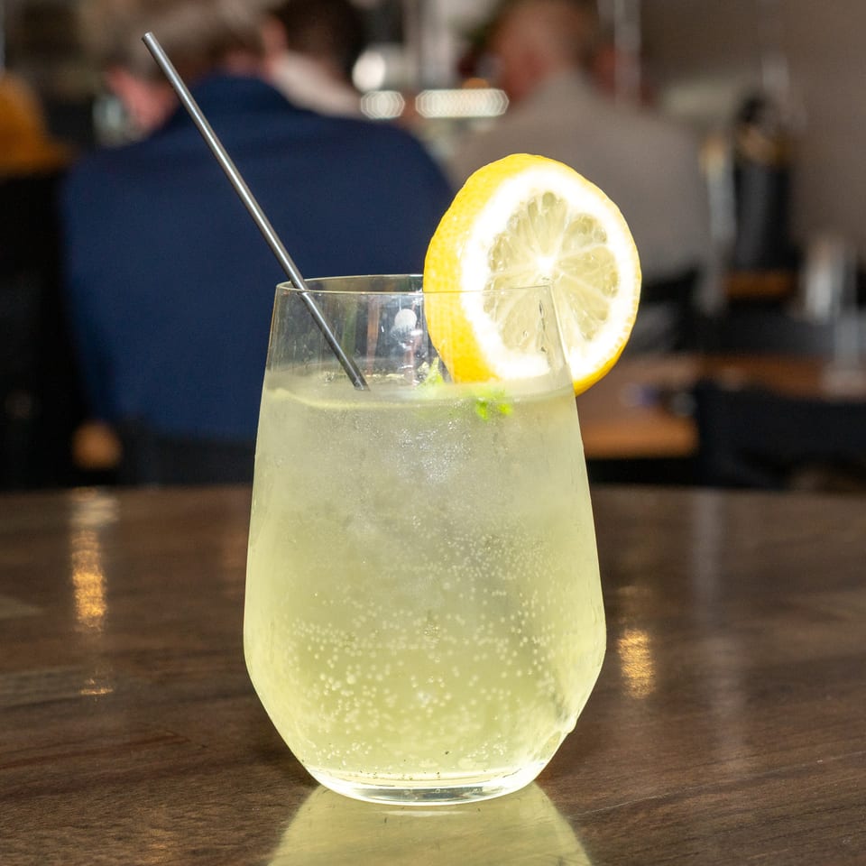 Sparkling lemonade with a lemon slice and metal straw on a wooden table, with blurred people in background.