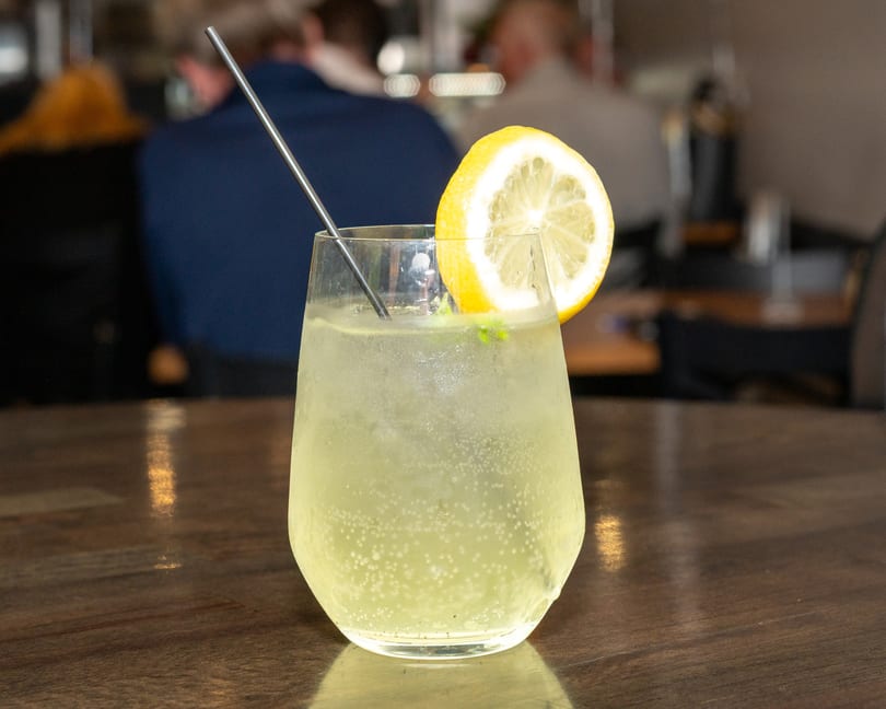 Sparkling lemonade with a lemon slice and metal straw on a wooden table, with blurred people in background.