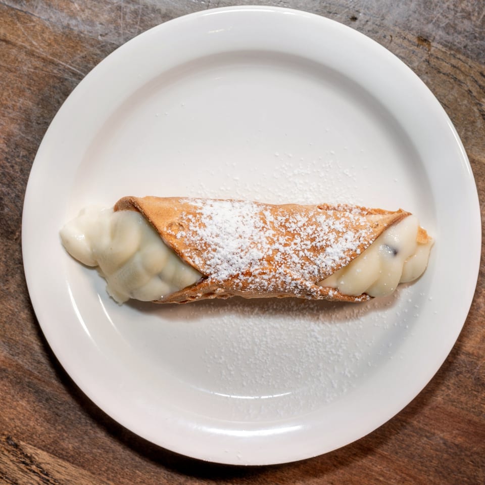 A single cannoli pastry, filled with cream and dusted with powdered sugar, on a white plate.