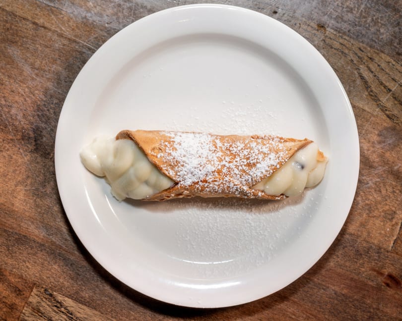 A single cannoli pastry, filled with cream and dusted with powdered sugar, on a white plate.