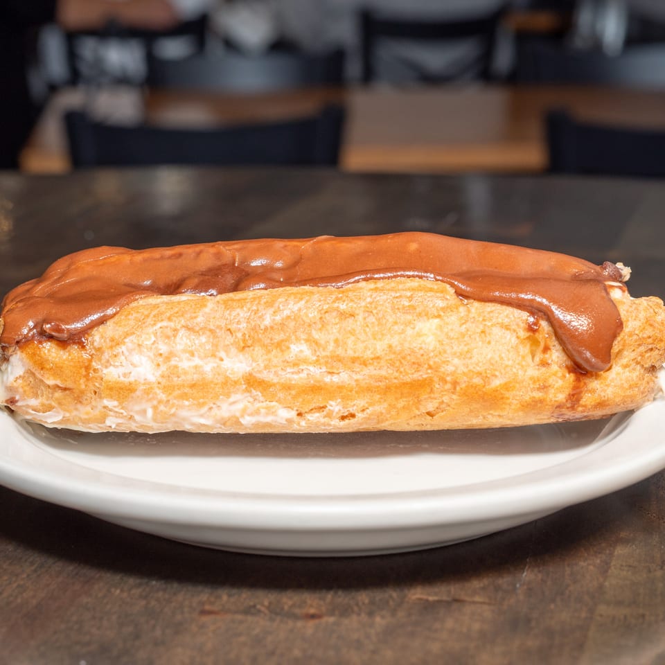 A chocolate-frosted eclair with white filling on a white plate, on a dark wooden table.