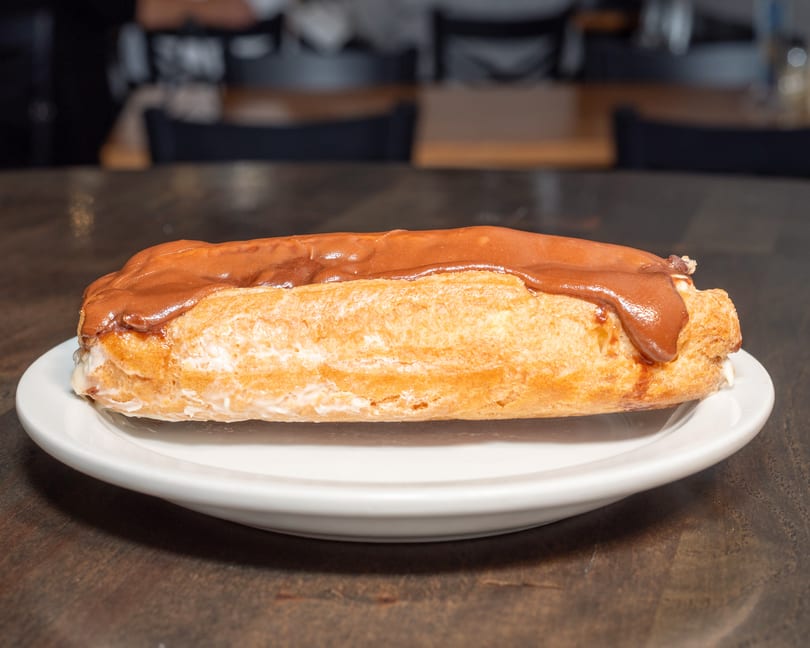 A chocolate-frosted eclair with white filling on a white plate, on a dark wooden table.
