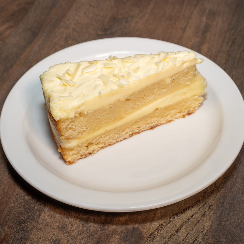 A slice of vanilla cake with white frosting and white chocolate shavings on a white plate.
