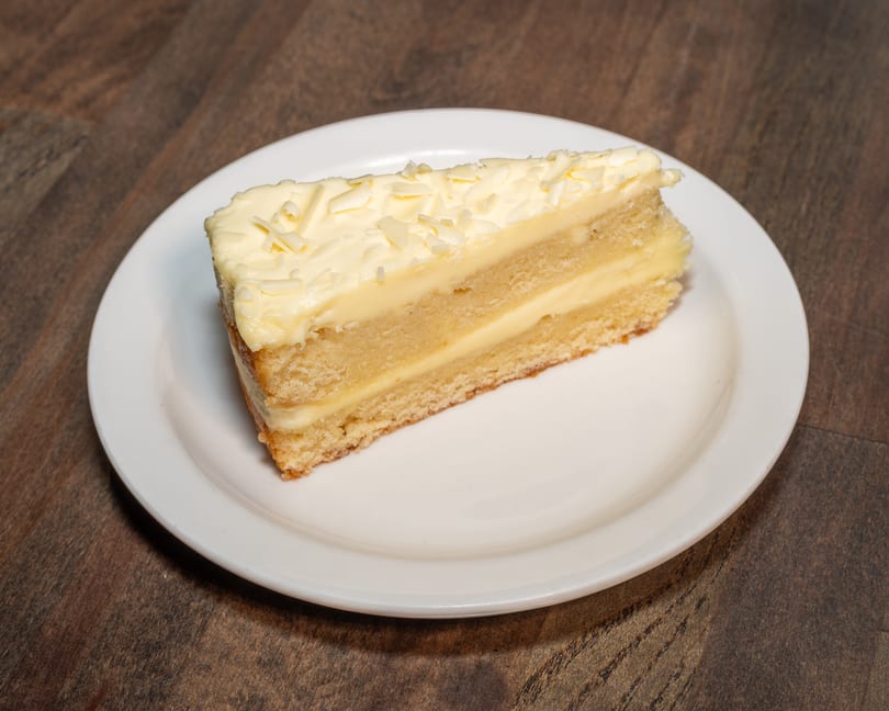 A slice of vanilla cake with white frosting and white chocolate shavings on a white plate.